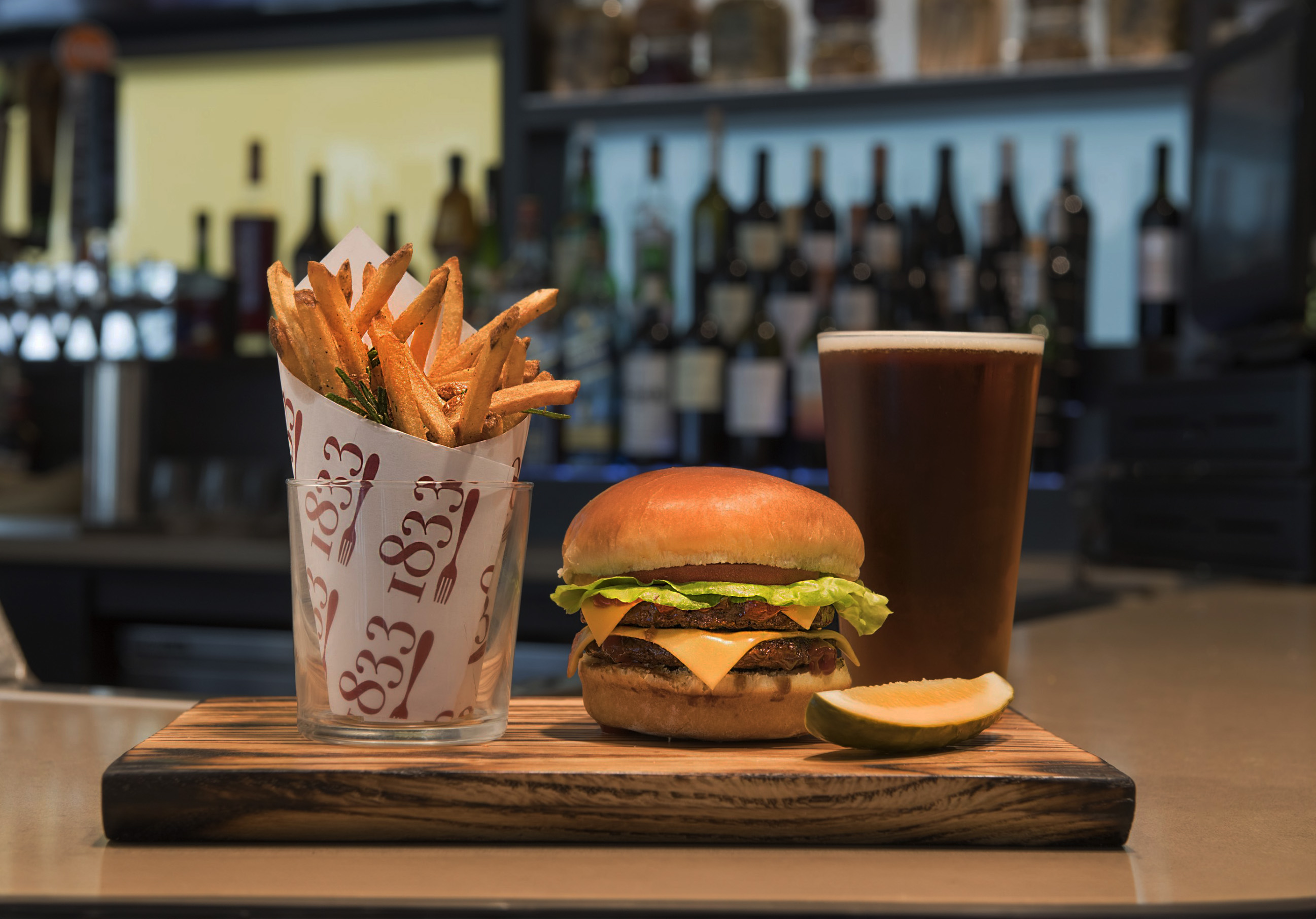 1833 burger and fries with a pickle and a glass of beer